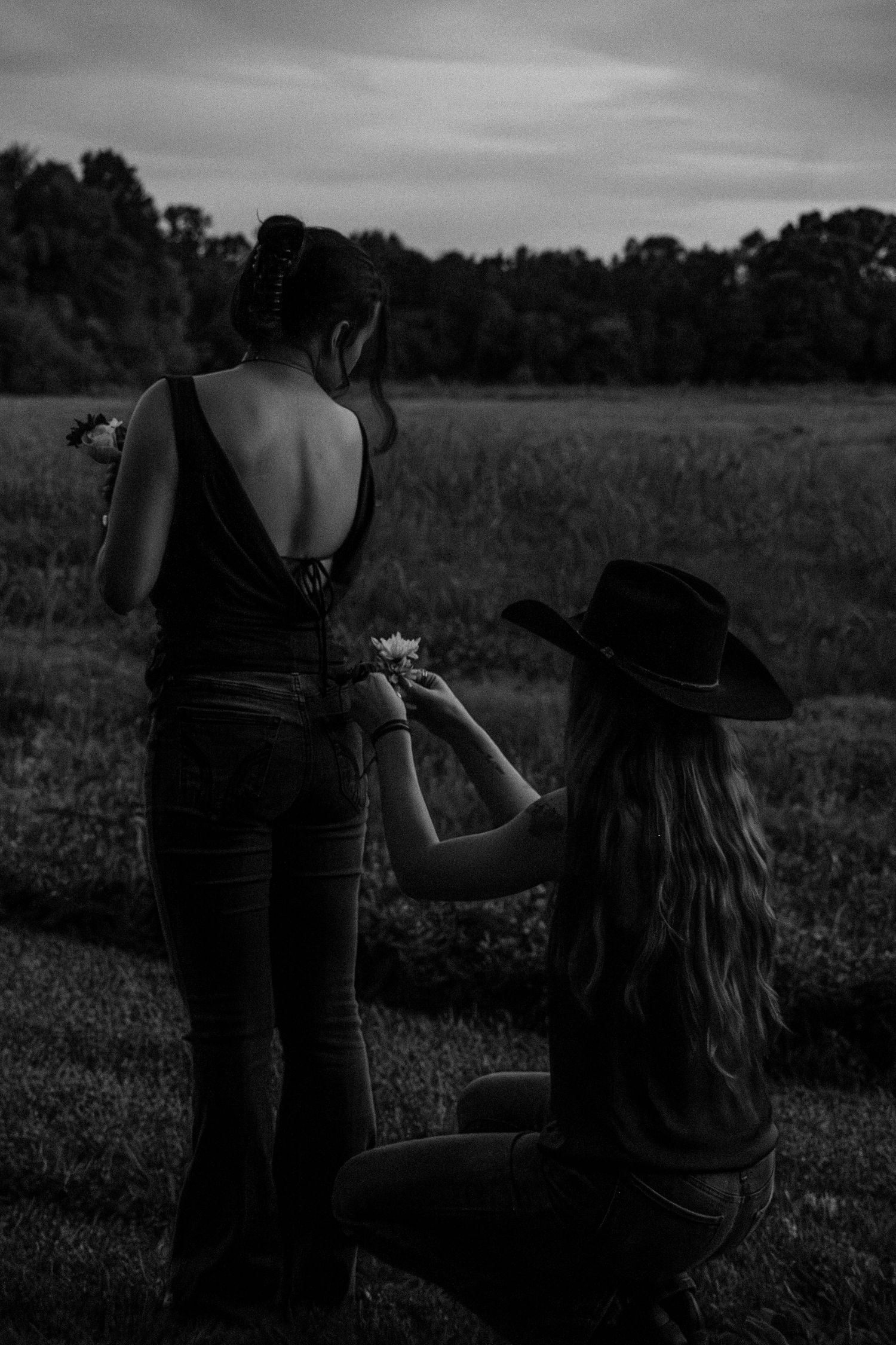 Two women stand in a field at dusk, one kneeling in a cowboy hat offering flowers, captured in black and white.