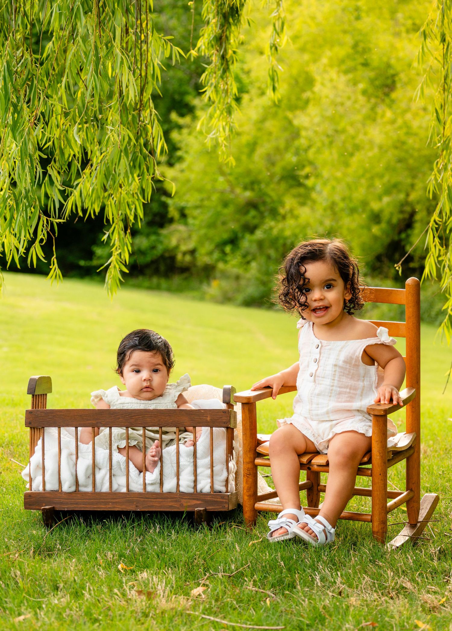 Two toddler girls pose outdoors, one in a wooden cradle and one on a rocking chair, under a weeping willow tree.