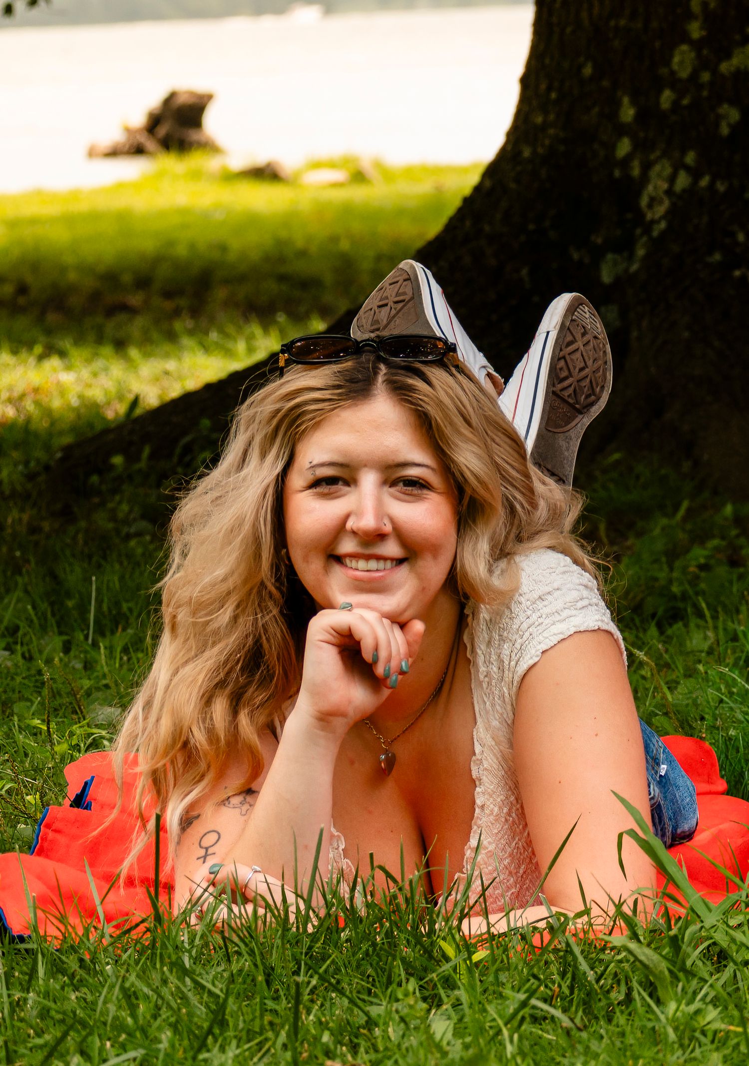 Smiling blonde woman lying on green grass under a tree, wearing a white top and red skirt, with sneakers visible.