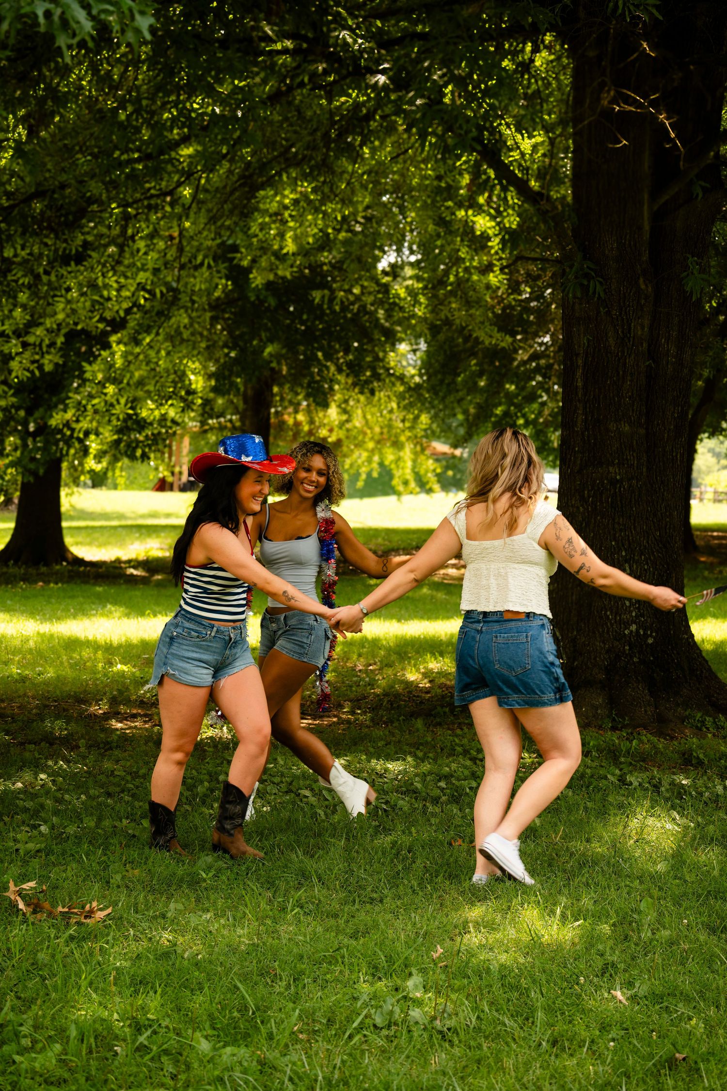 Three women in denim shorts dance together on green grass in a sunny park setting.