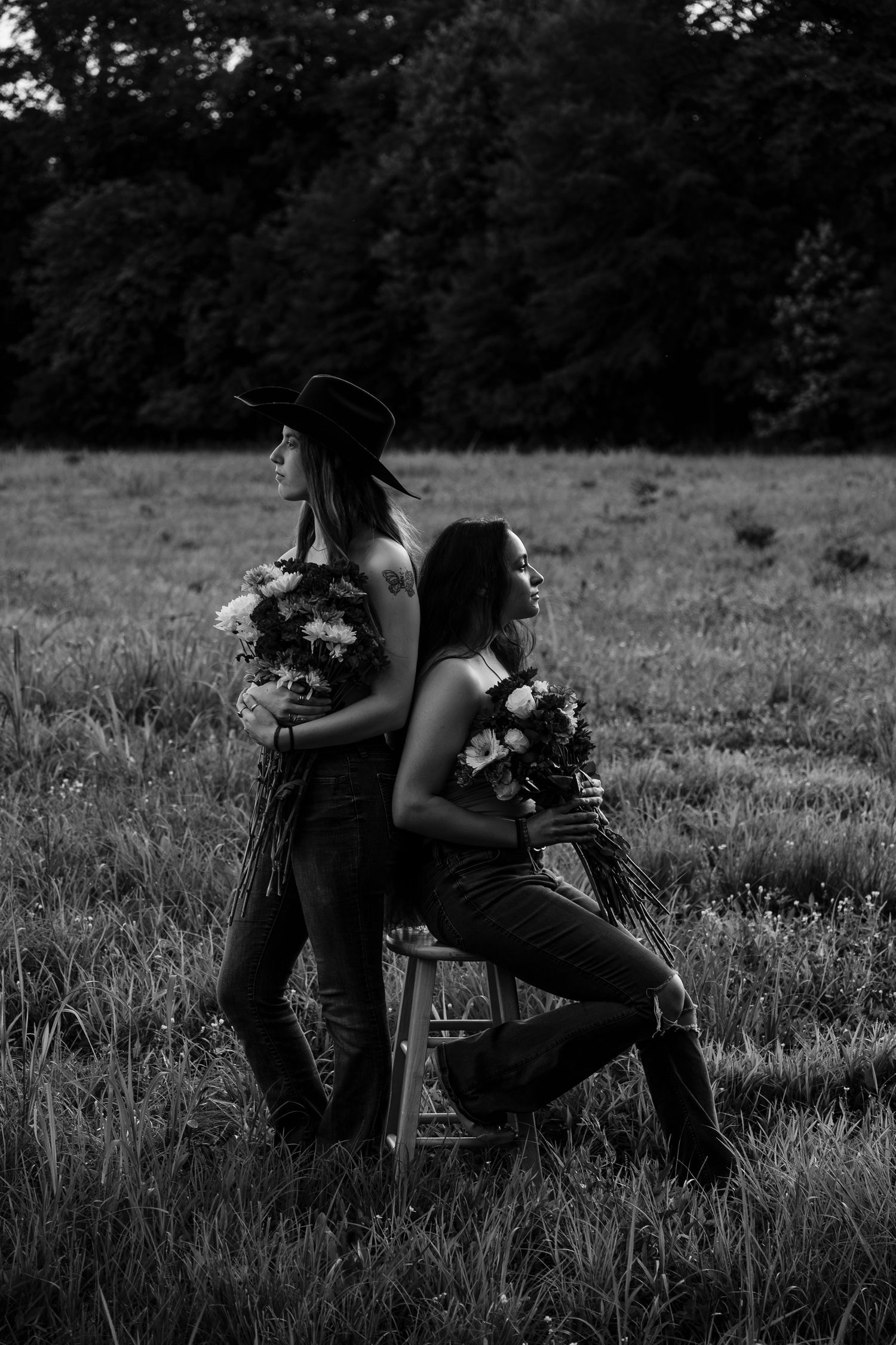 Two women pose back-to-back in a field holding flower bouquets, one wearing a cowboy hat, in black and white photography.