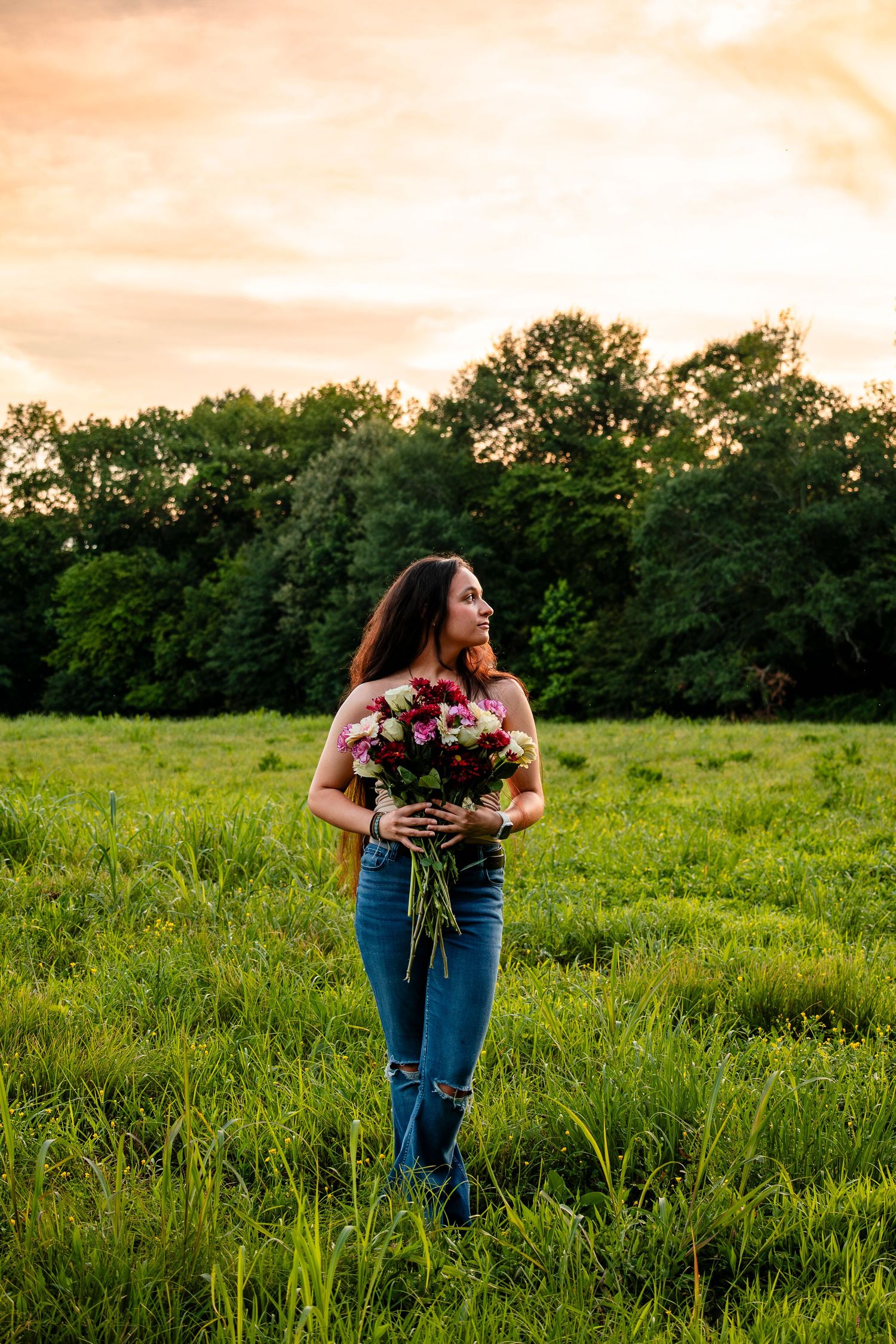 Woman holding colorful flower bouquet standing in green field at golden hour sunset with trees in background.