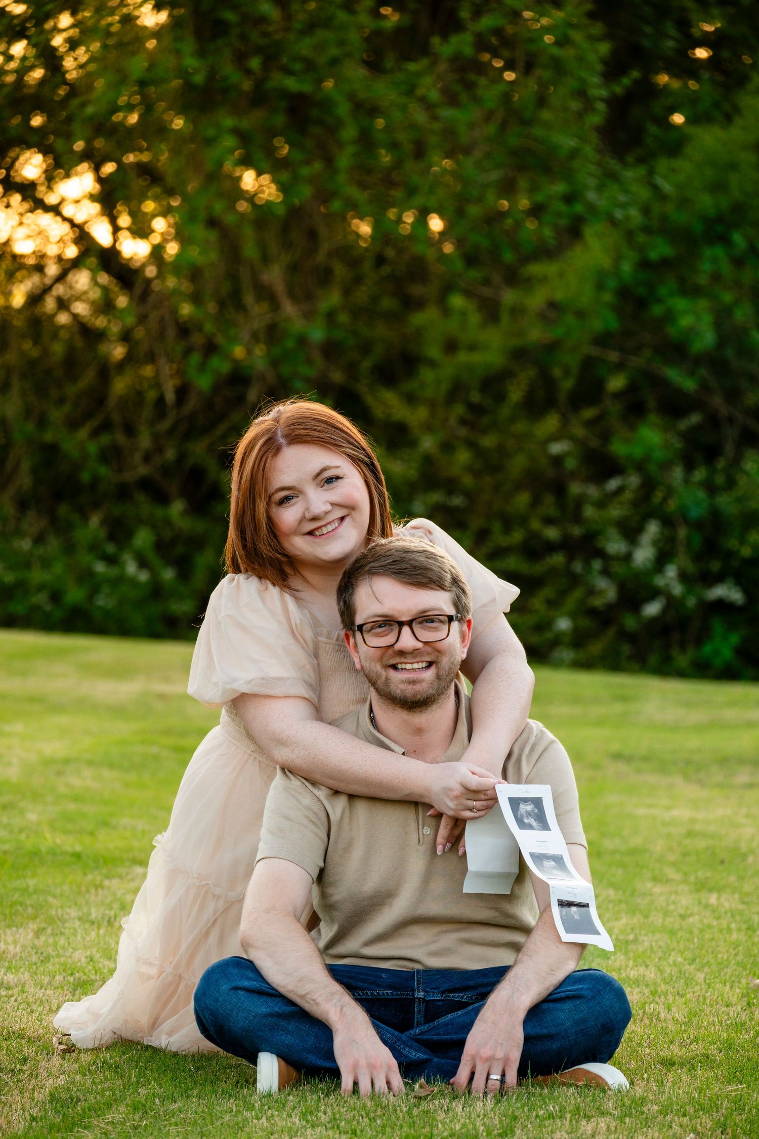Joyful redhead woman embracing seated husband holding ultrasound photos on lush green lawn at dusk.