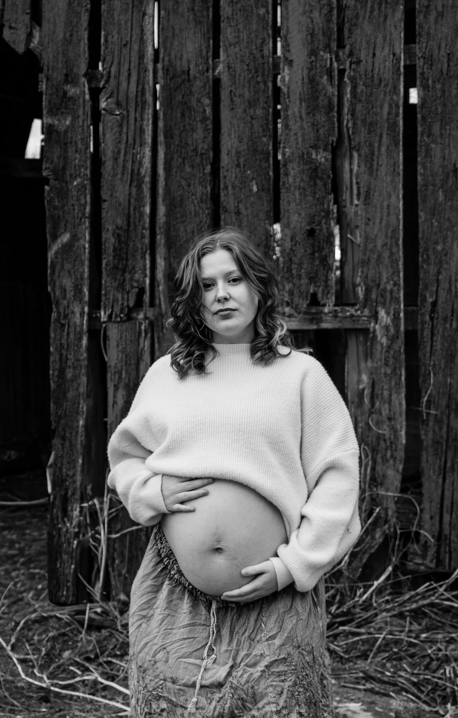 Pregnant woman in white sweater holds baby bump outdoors near rustic wooden barn wall, black and white maternity photo.