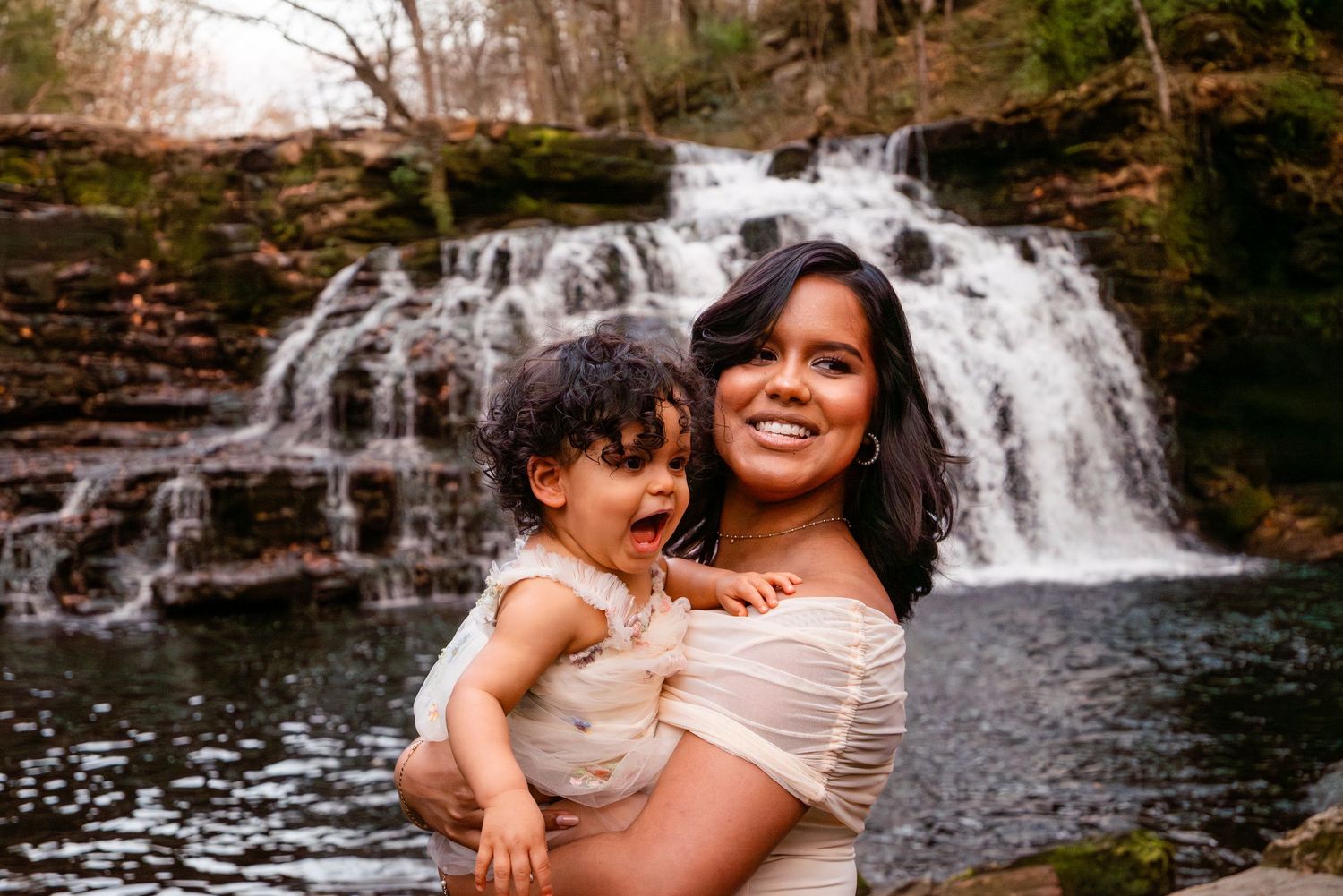 Smiling pregnant woman holds laughing toddler in front of a beautiful cascading waterfall outdoors.