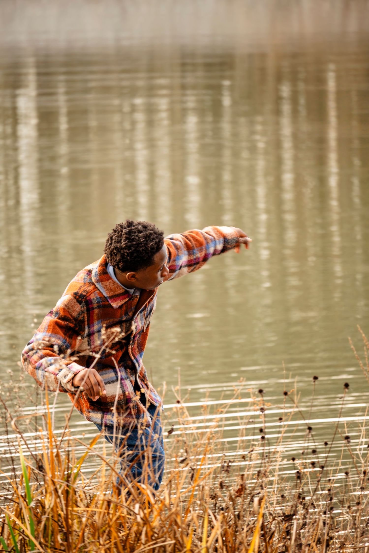 A young Black boy skips stones at the edge of a reflective lake wearing a plaid flannel shirt.