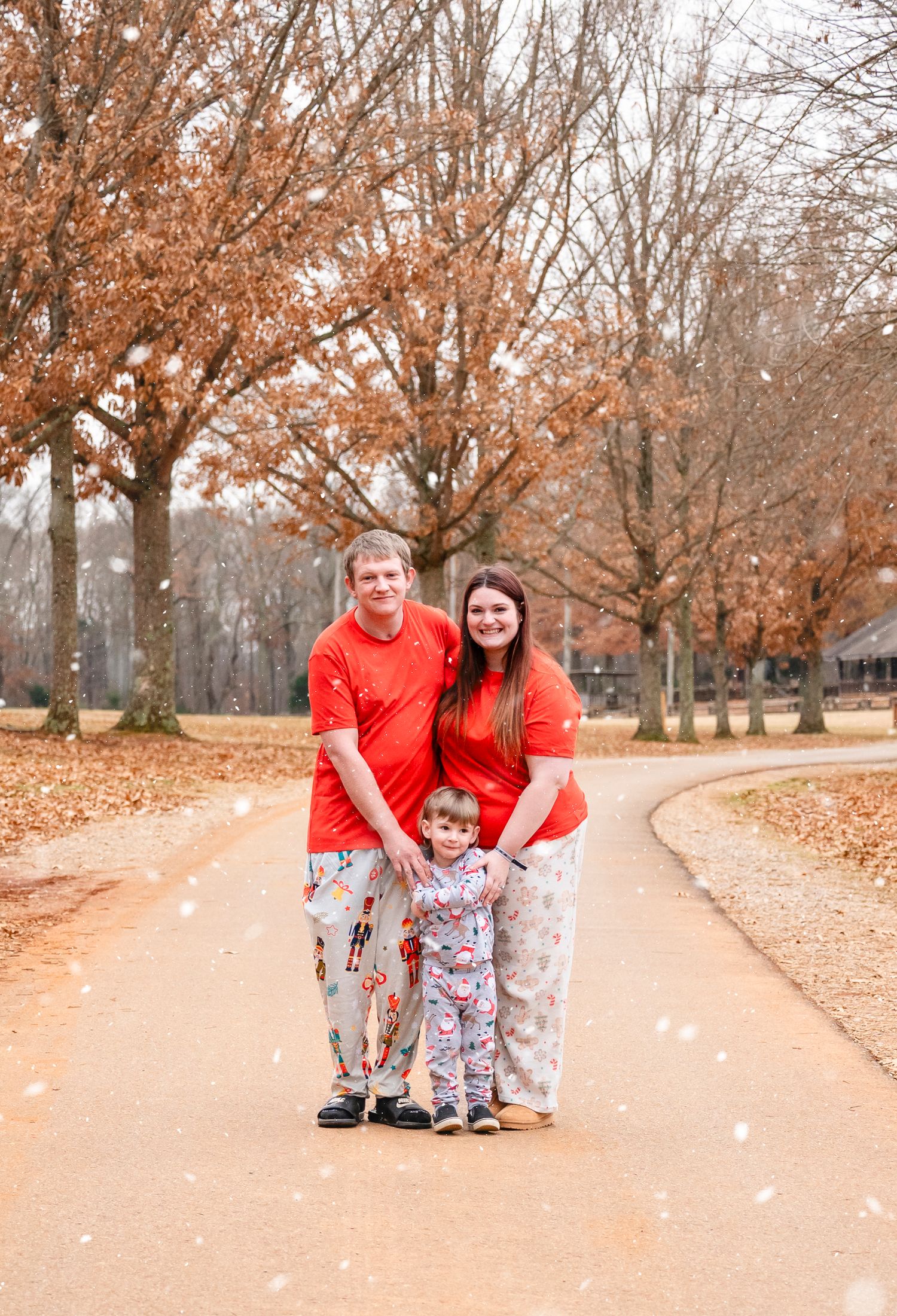 Family of three in matching red Christmas pajamas poses on a tree-lined path with artificial snow.