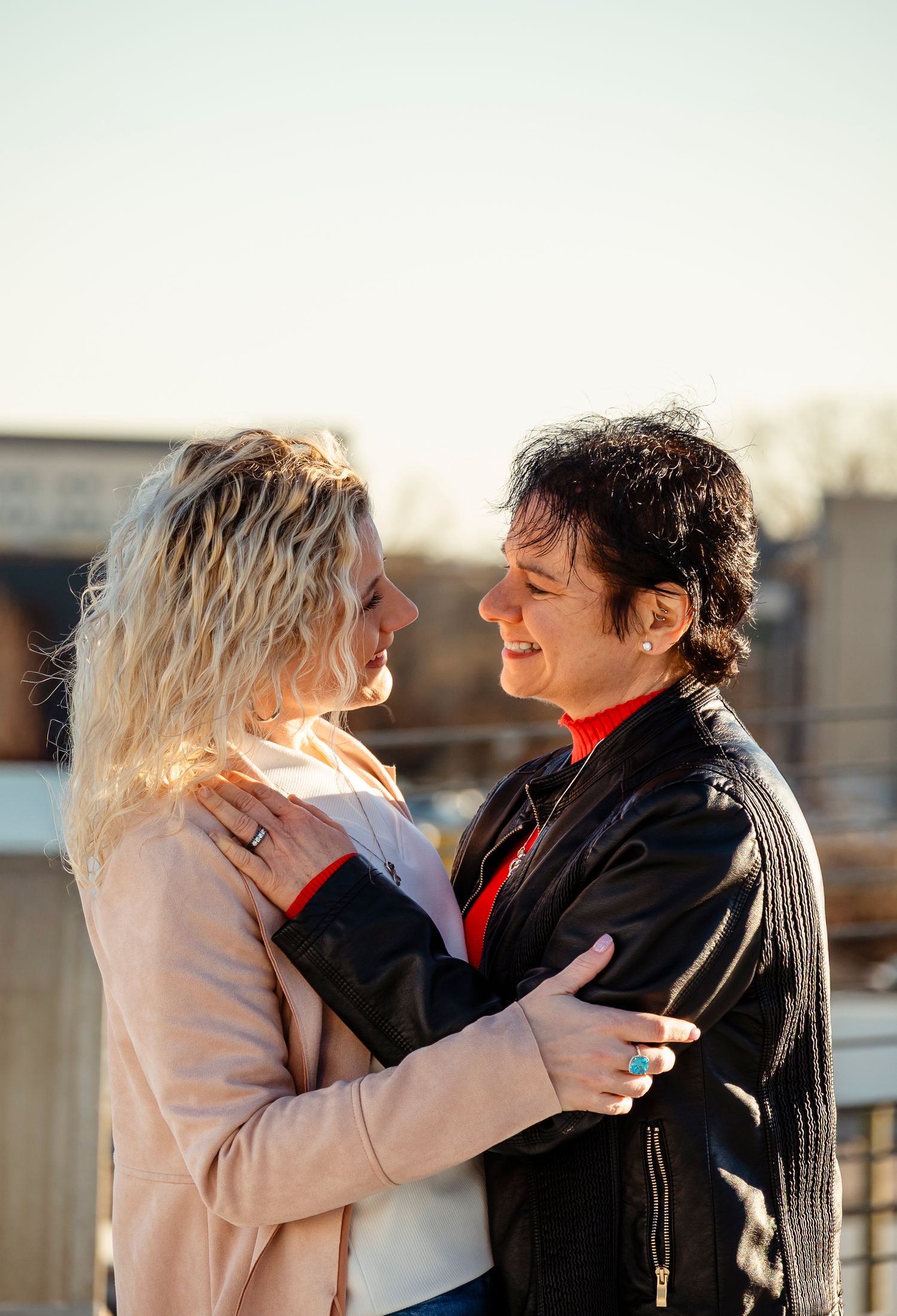 Two women smiling and embracing outdoors in warm sunlight during an engagement photo session.