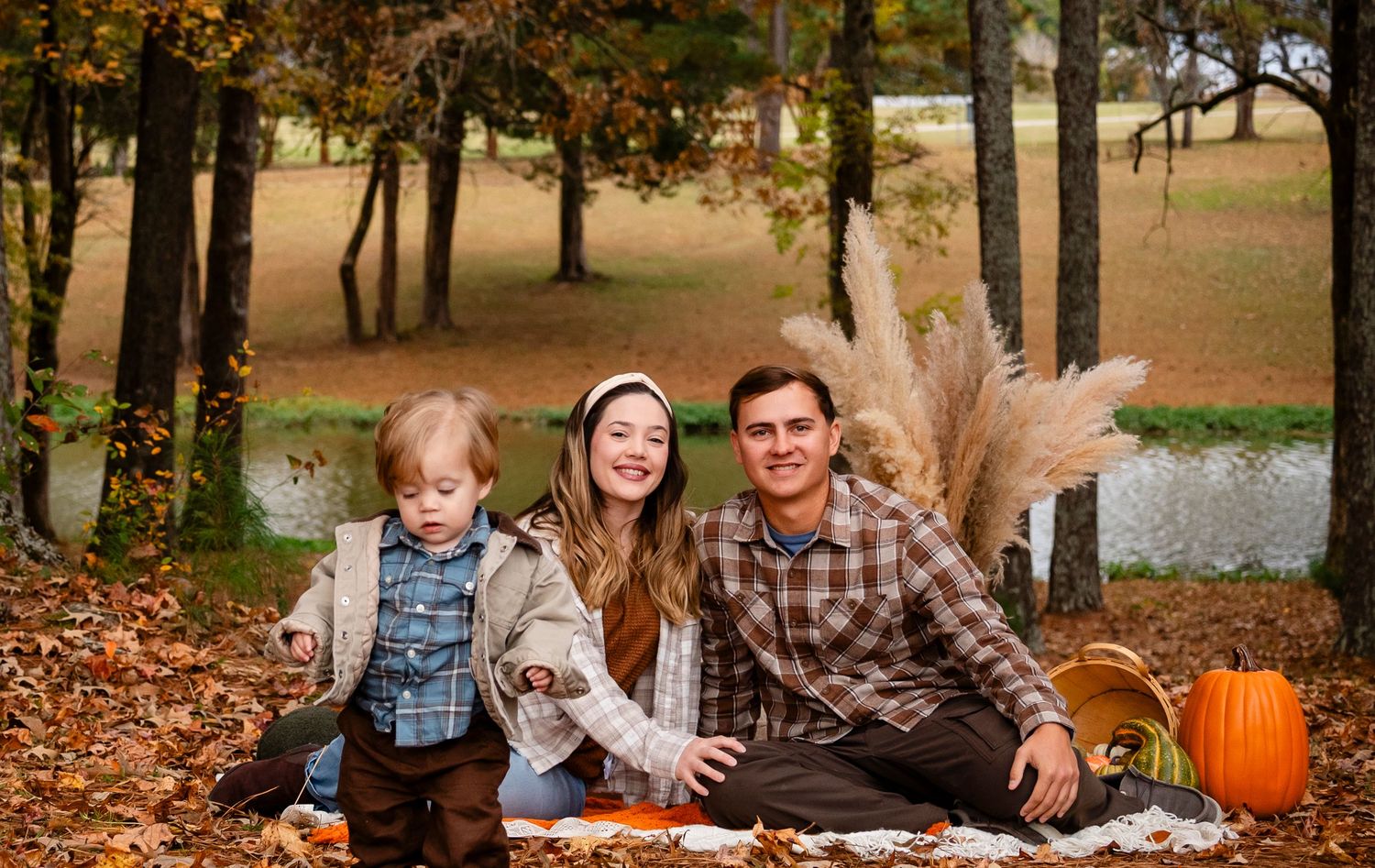 Family of three posing outdoors on fall leaves with pumpkins and pampas grass decor.