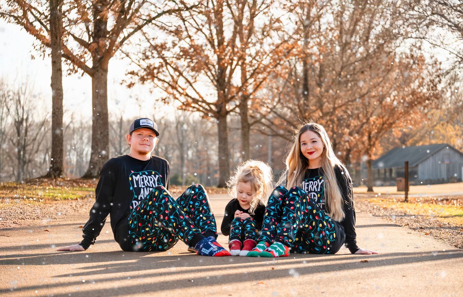 Three siblings in matching black Christmas light pajamas sitting on sunlit path in autumn park.