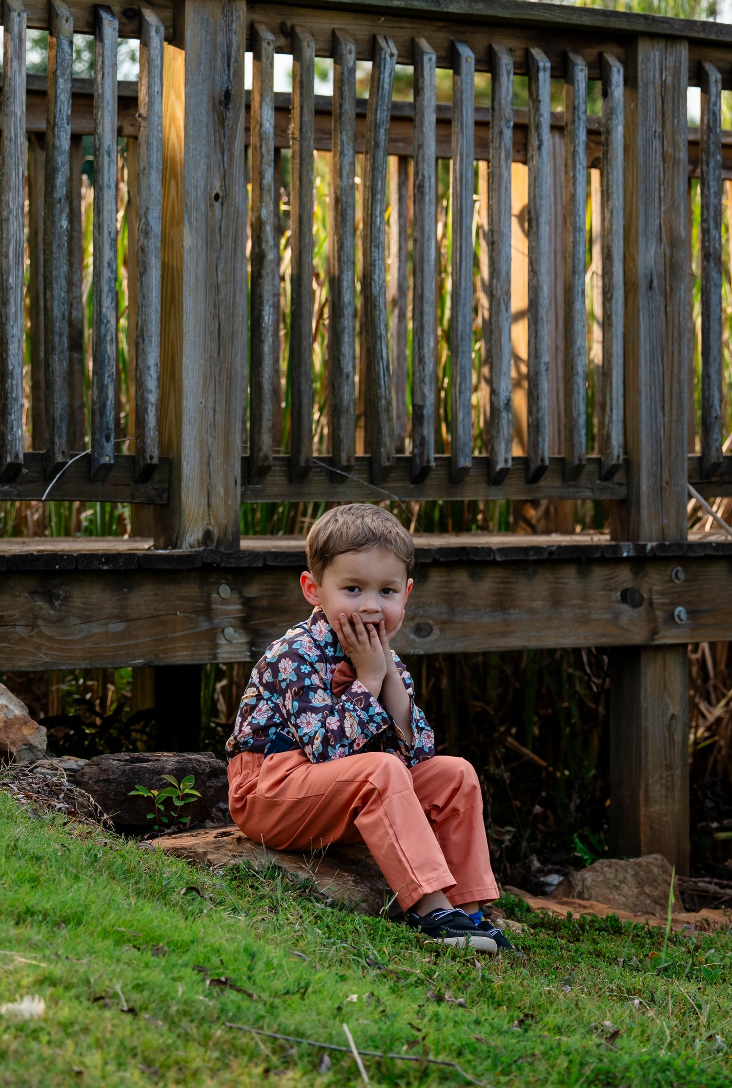 Young boy in orange pants and floral shirt sitting on rocks near a rustic wooden fence outdoors.