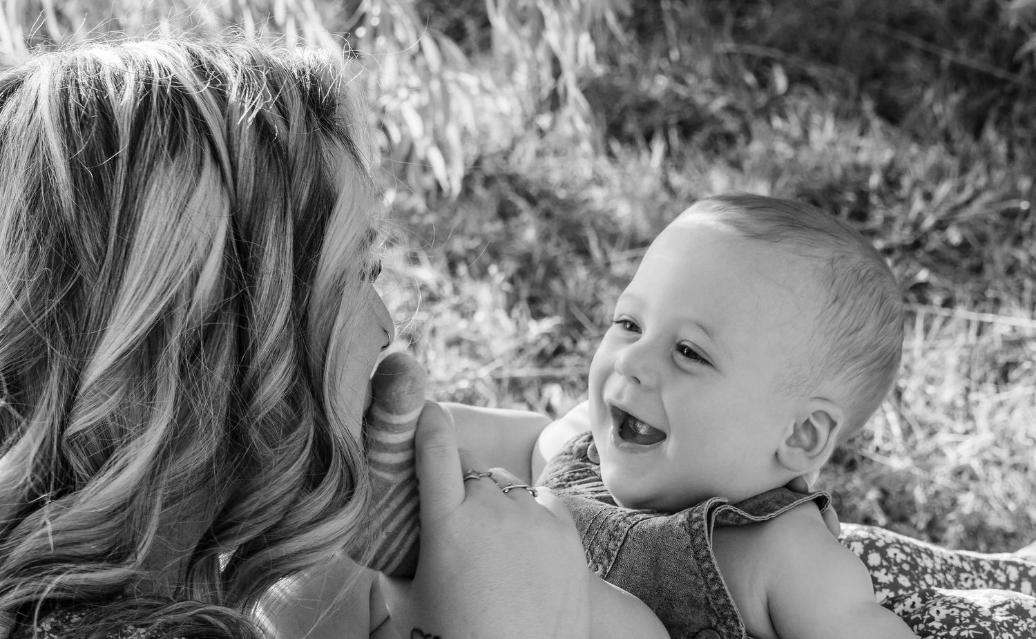 Black and white photo of a joyful baby laughing while being held by a smiling blonde woman outdoors.