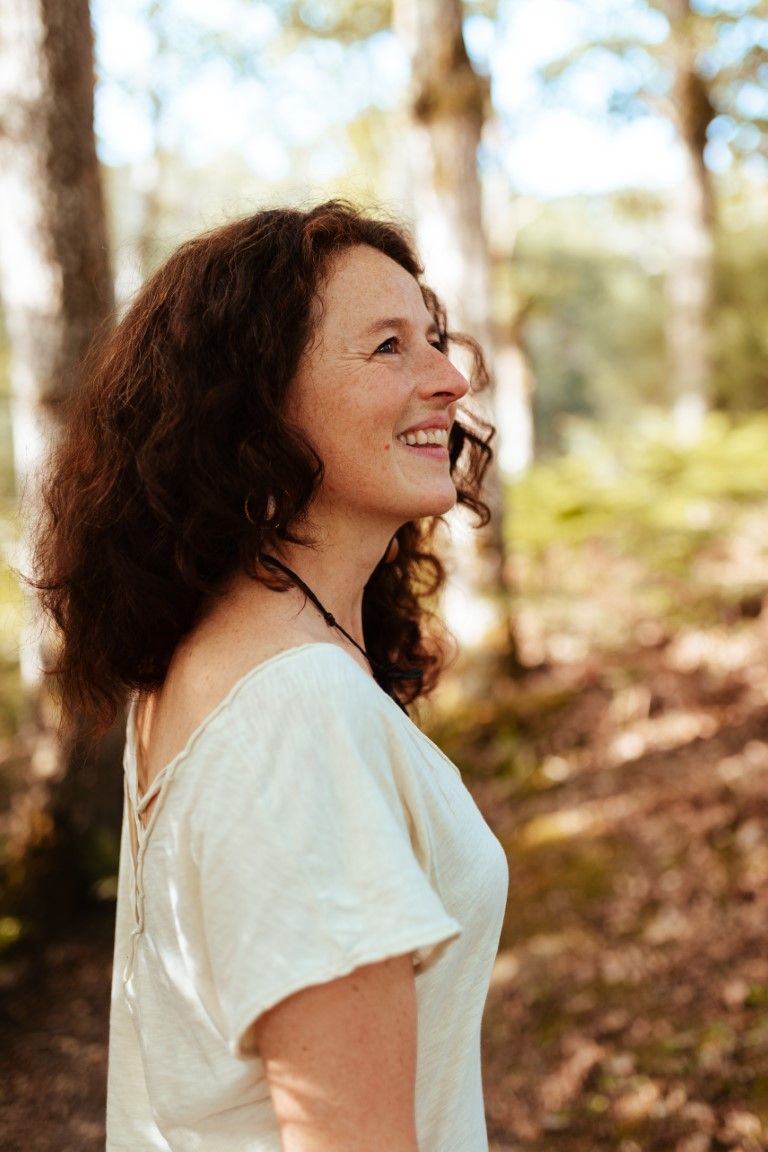 A smiling woman with curly dark hair wearing a white top stands outdoors in a sunlit forest.