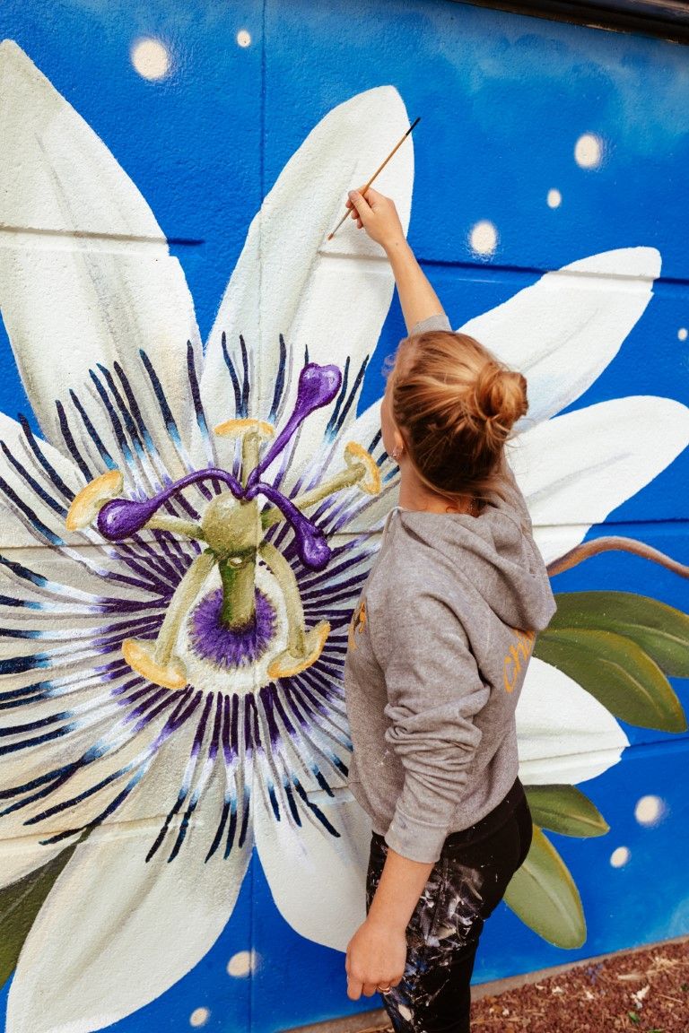 Female artist painting detailed passionflower mural with brush on blue outdoor wall.