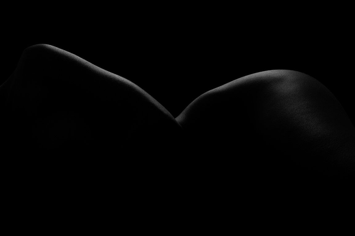 Black and white fine art bodyscape photograph highlighting curves and contours of human form against dark background.