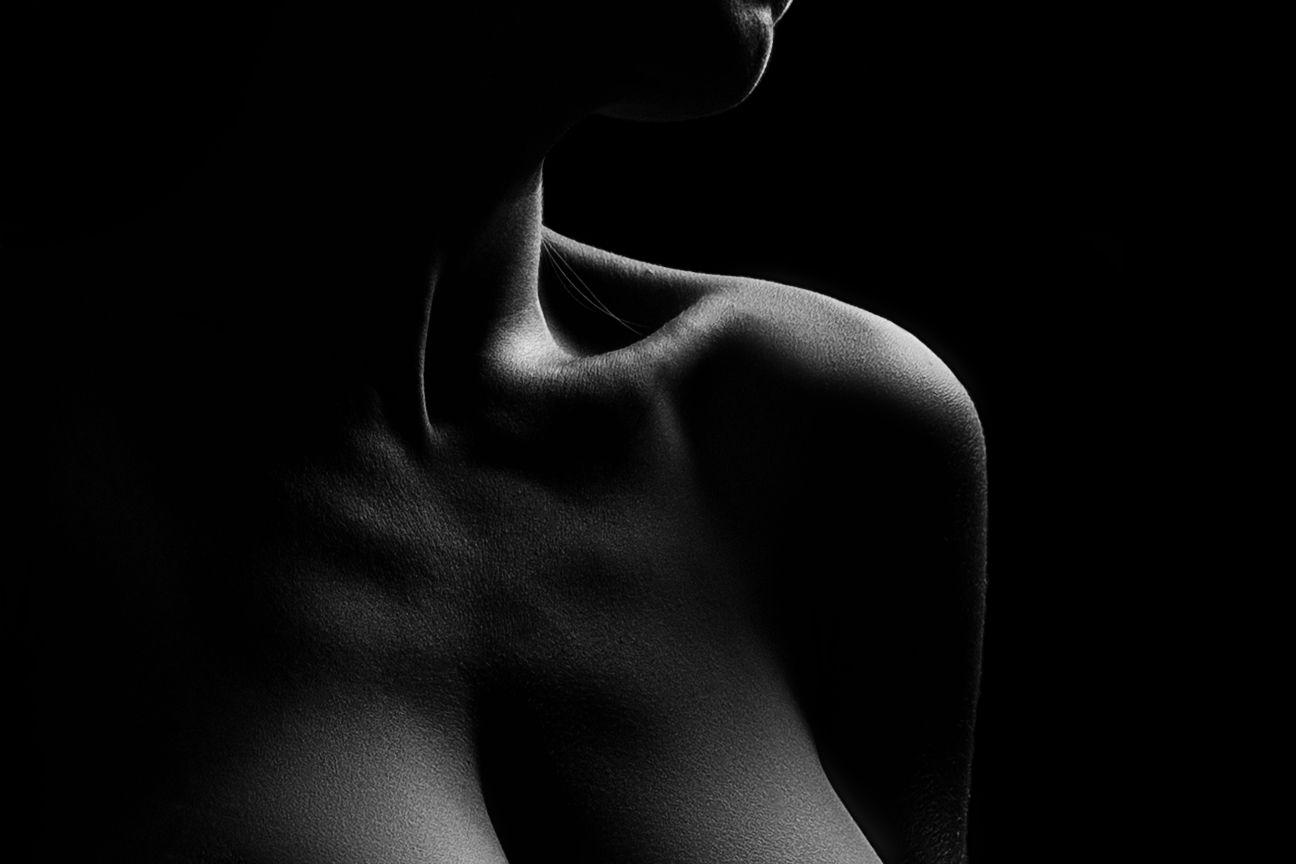 Black and white artistic nude photography highlighting elegant shoulder and collarbone contours against dark background.