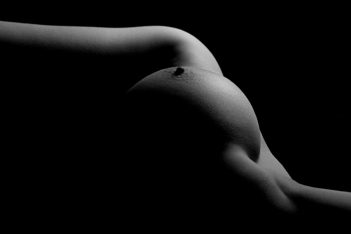 Black and white artistic nude bodyscape photograph highlighting elegant curves against a dark background.