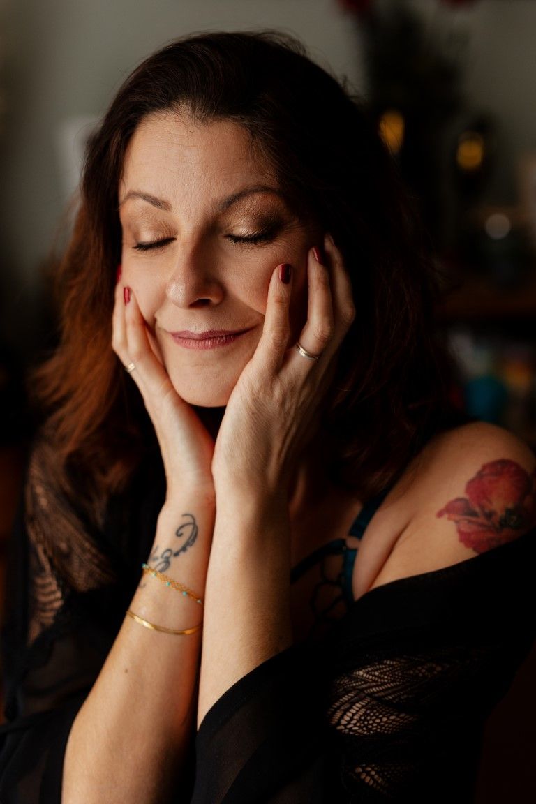 Une femme brune souriante, tatouée, se tient doucement le visage entre ses mains, les yeux fermés.
