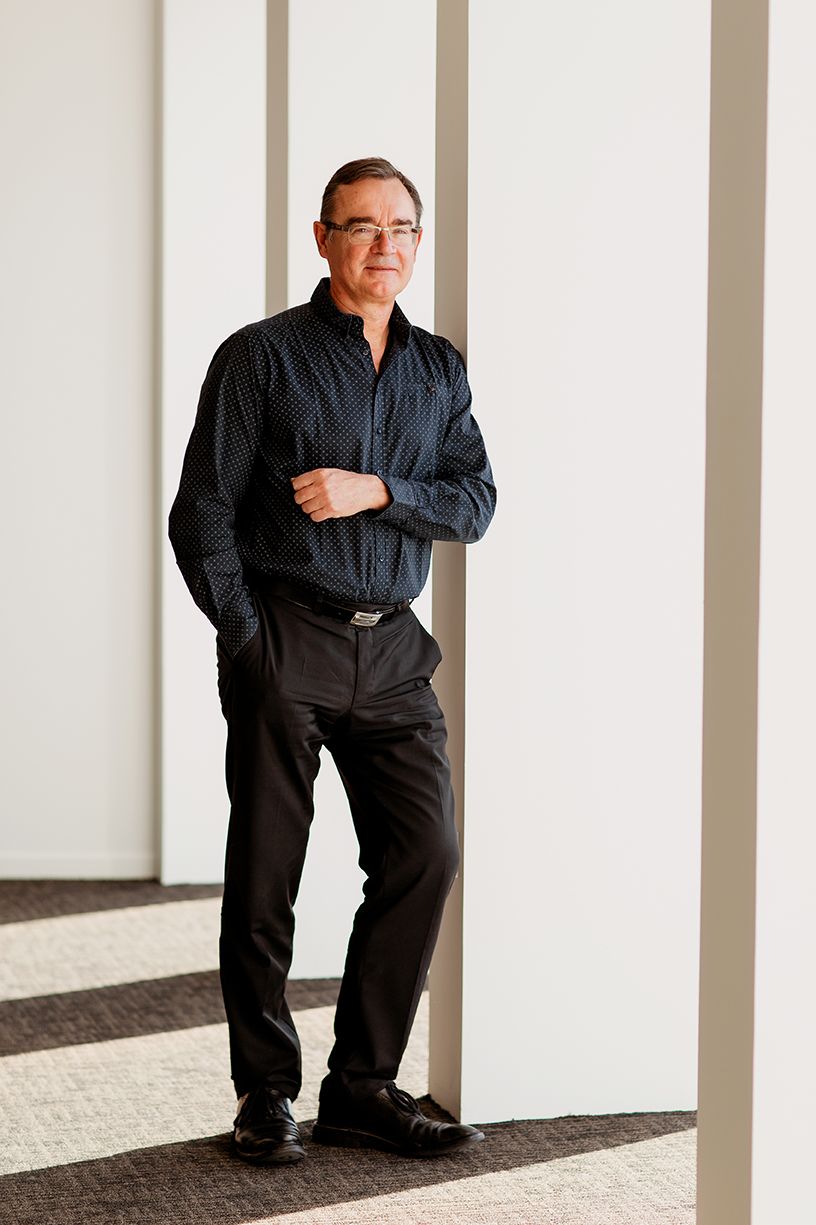 Middle-aged man in black patterned shirt and trousers leaning against white pillar in modern office.