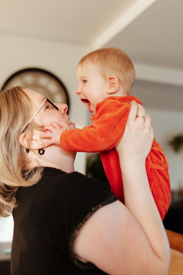 Un bébé joyeux vêtu d'une tenue orange est soulevé par sa mère souriante qui porte des lunettes.