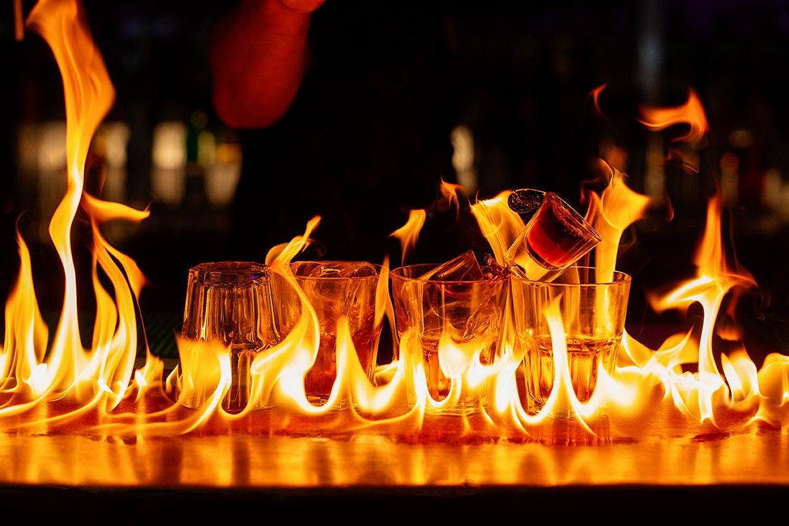 Row of shot glasses engulfed in dramatic orange flames on a dark bar counter.