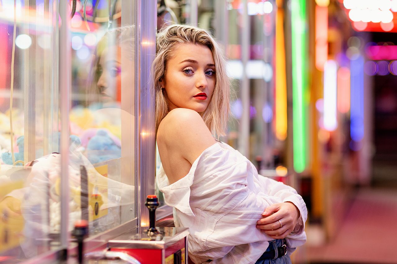 Femme blonde en haut blanc à épaules dénudées posant dans une salle de jeux colorée.