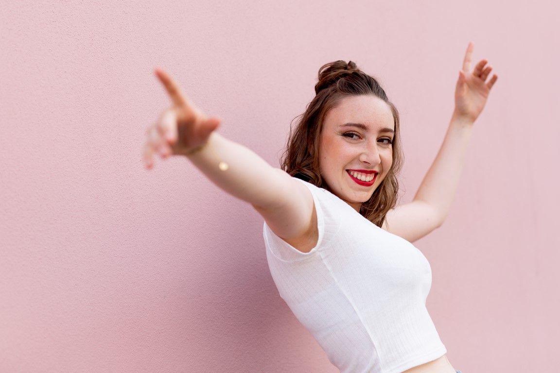 Jeune femme en crop top blanc souriant et pointant du doigt sur fond rose.