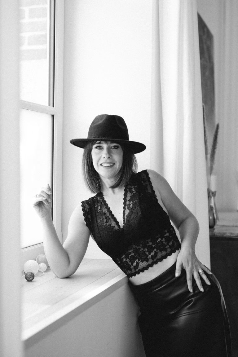 Black and white photo of brunette woman in black lace top and hat posing by window.