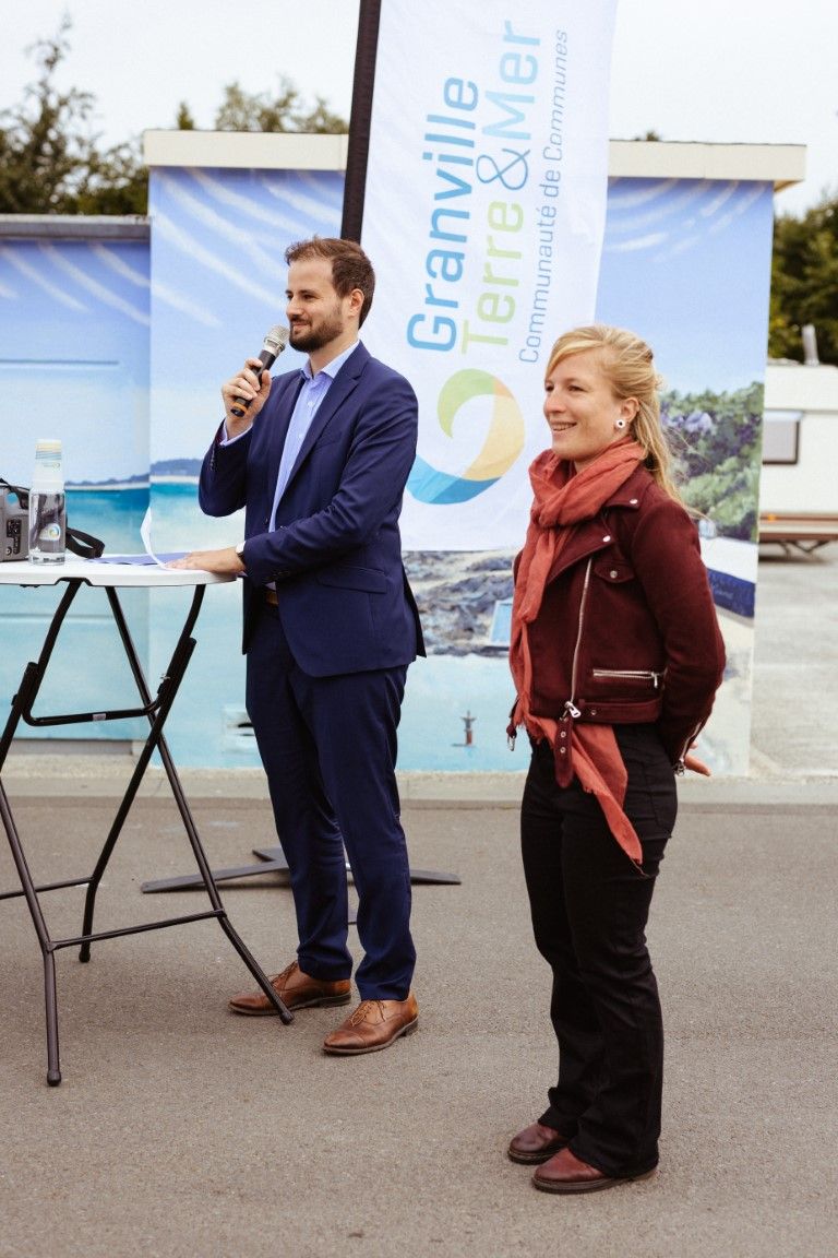 A man in a blue suit speaks at a microphone beside a woman in a red jacket at a Granville Terre & Mer community event.