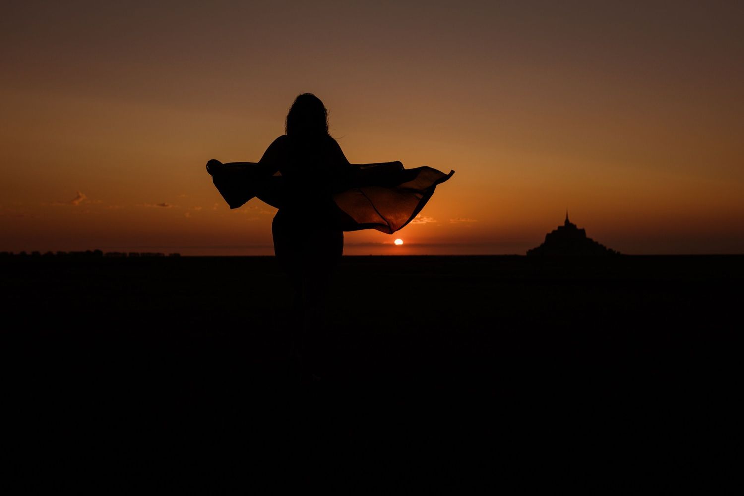 Silhouette of a person with flowing cape at sunset, with a distant landmark on the horizon.