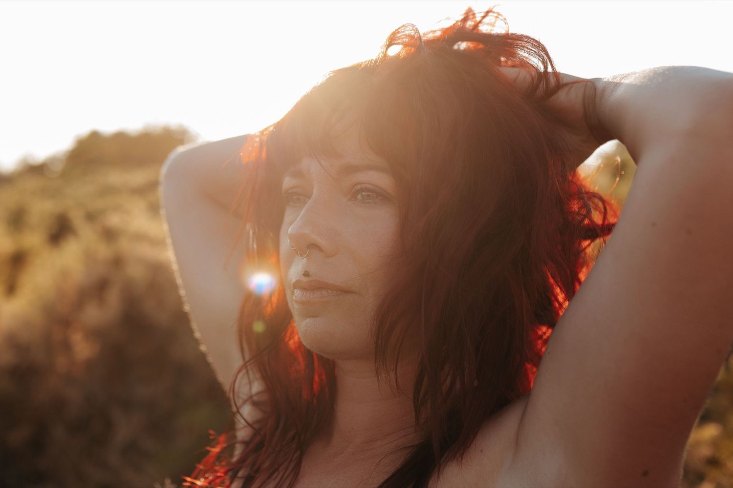 Woman with red hair poses outdoors in golden sunset light, hands raised behind head, with warm lens flare effect.