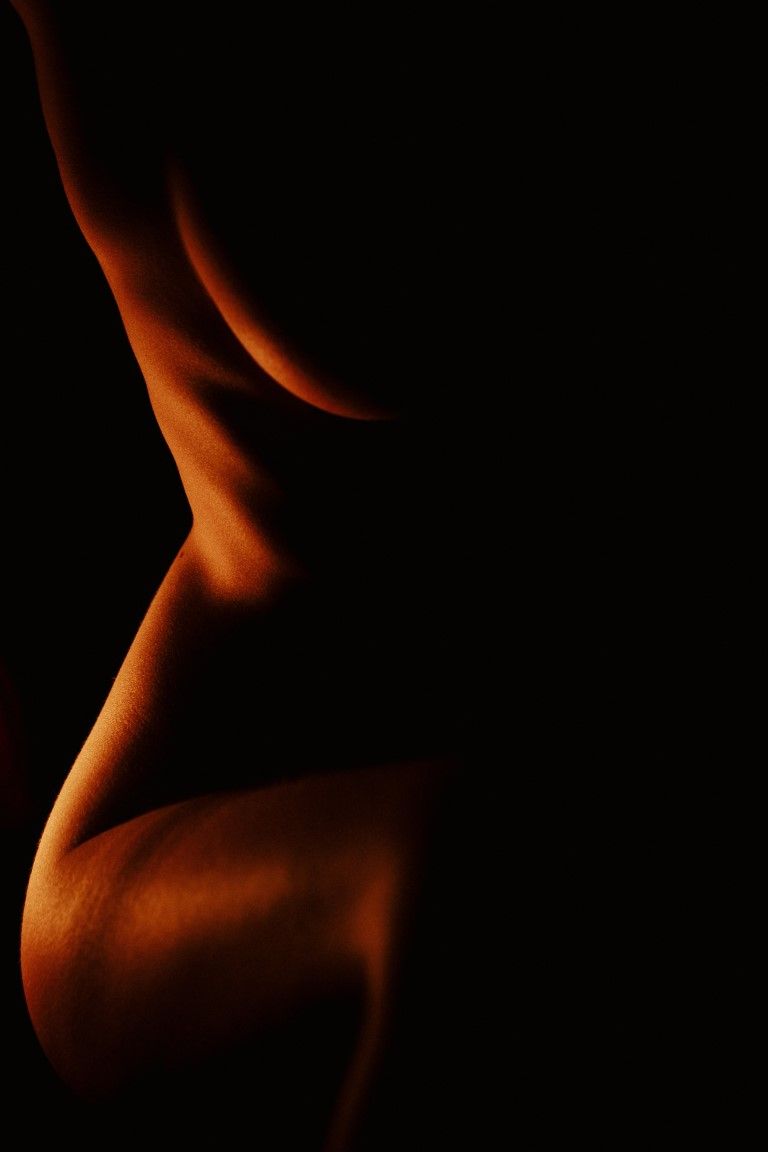 Silhouette nude discrète et spectaculaire, illuminée par une chaude lumière orangée qui met en valeur les courbes féminines sur fond noir.