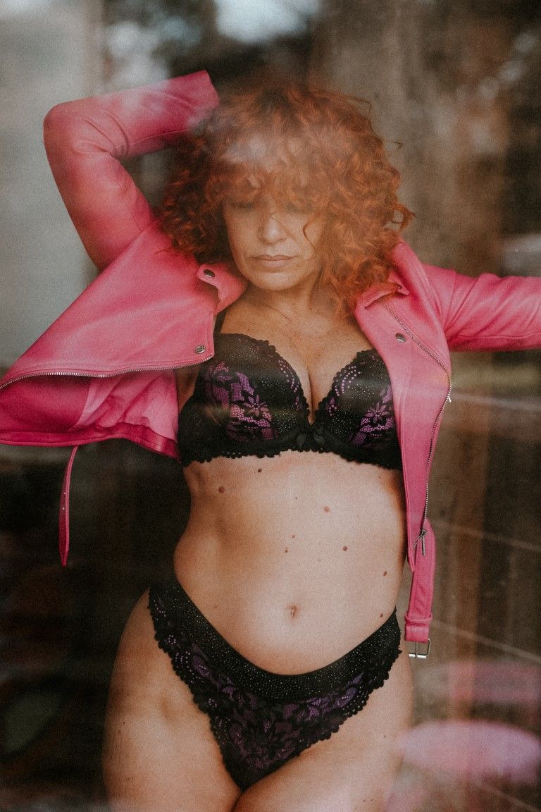 Redhead woman in black lace lingerie and pink leather jacket poses behind glass window.