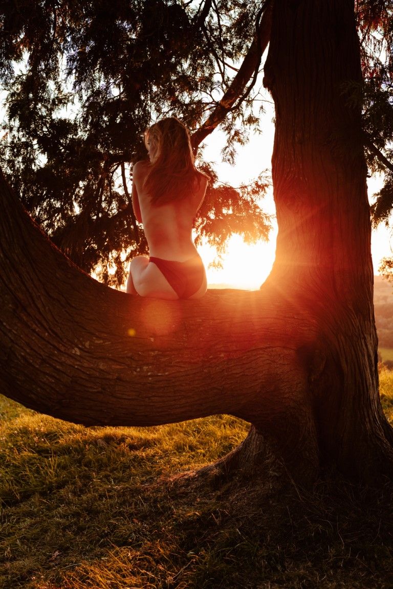 Femme en maillot de bain blanc perchée sur un tronc d'arbre courbé, se détachant en silhouette sur la lumière éclatante du coucher de soleil
