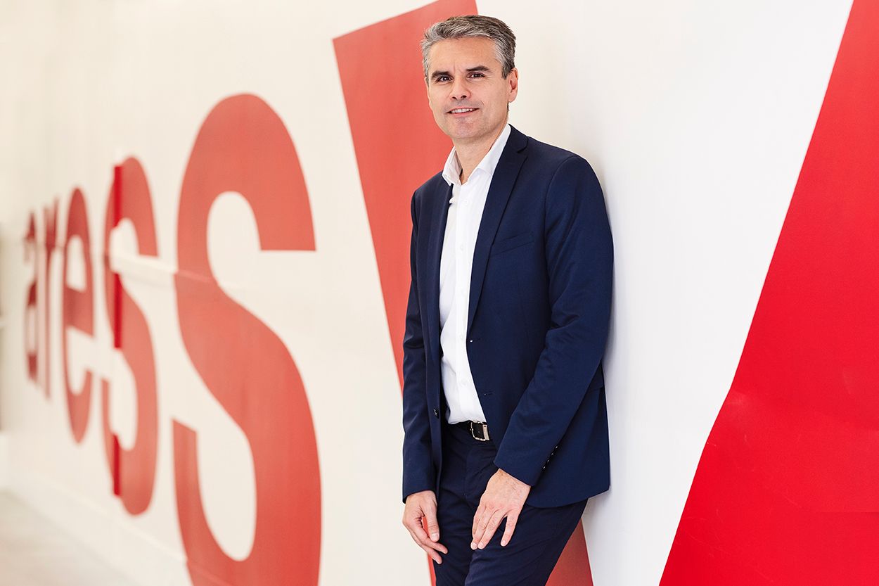 Businessman in navy blazer leaning against red and white branded wall smiling.