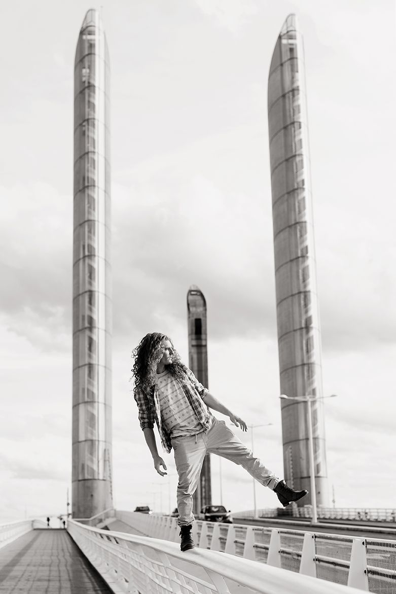 Photo en noir et blanc d'une personne aux cheveux bouclés adoptant une pose dynamique sur un pont à hauts piliers.