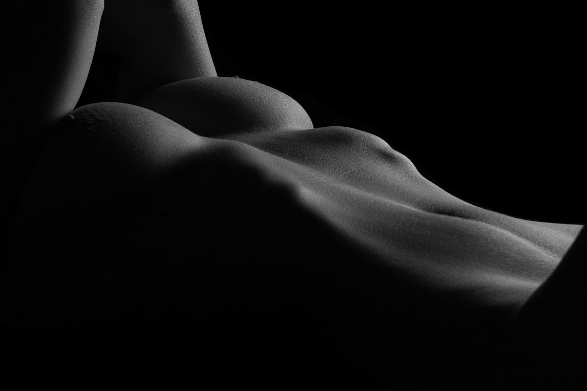 Black and white artistic nude bodyscape photograph highlighting curves with dramatic chiaroscuro lighting on dark background.