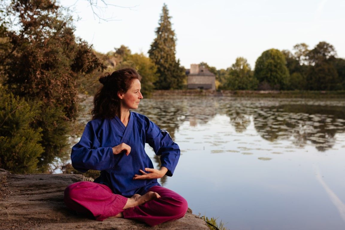 Woman meditating in lotus pose by a serene lake, wearing blue top and pink pants, practicing tai chi outdoors.