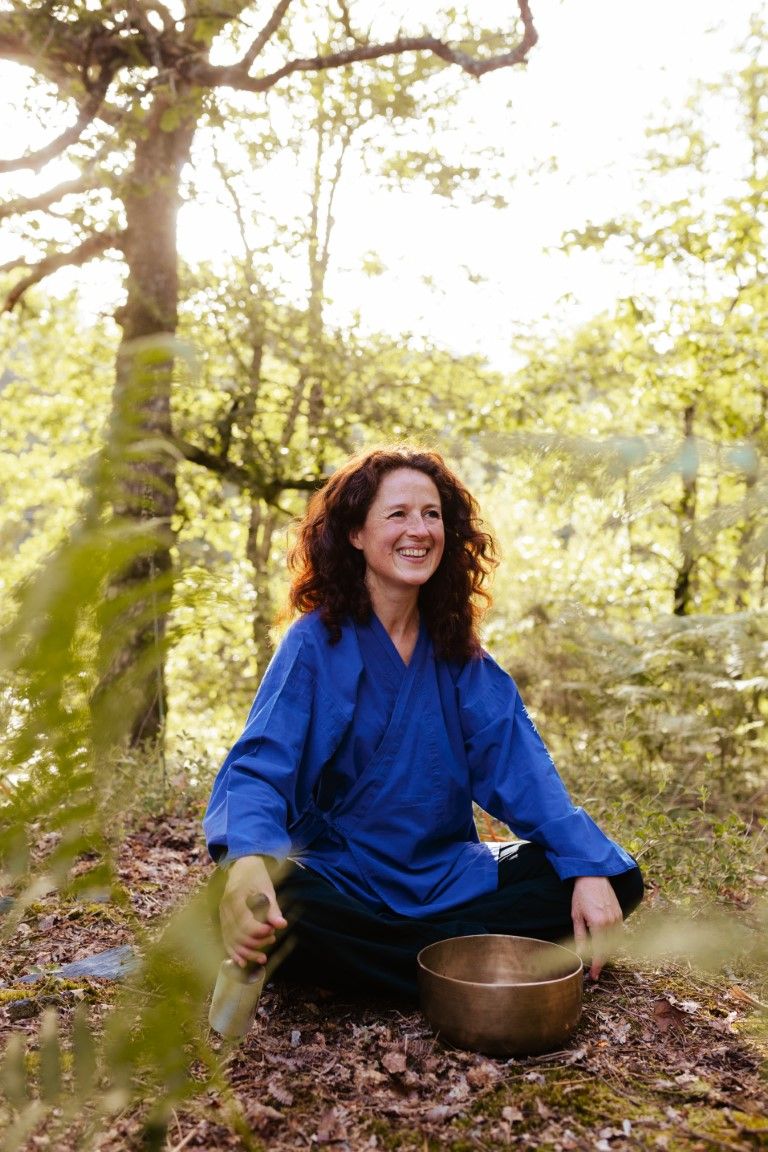 Smiling woman with red hair in blue kimono sits cross-legged in forest with singing bowl, enjoying outdoor meditation.