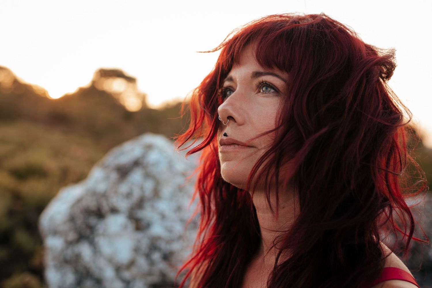 A woman with vibrant red wavy hair gazes upward outdoors at golden hour with rocks and hills in the background.