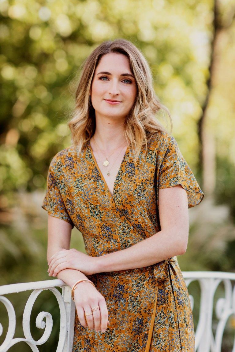 portrait Une femme blonde souriante, vêtue d'une robe portefeuille à fleurs moutarde, est appuyée contre une clôture blanche à l'extérieur.