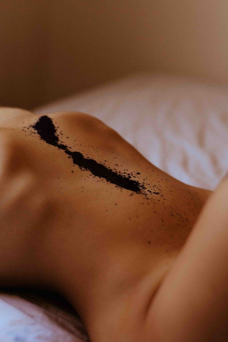 Artistic nude body photography with black powder scattered across bare lower back on white sheets.