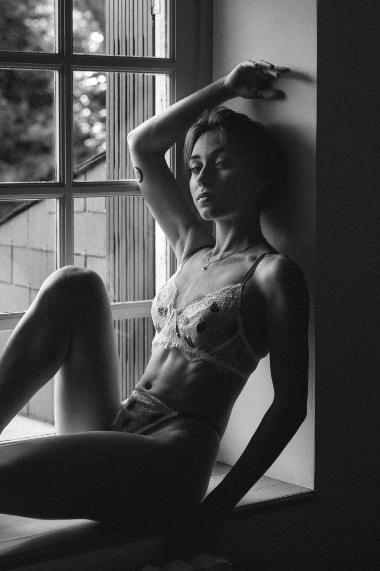 Black and white boudoir portrait of a slender woman in lace lingerie sitting in a window.