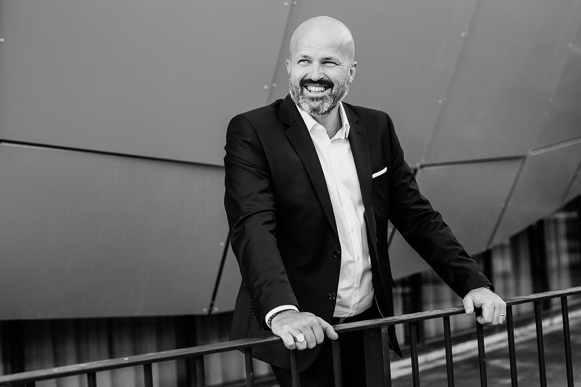 Smiling bald man in black suit leaning on railing in black and white portrait.