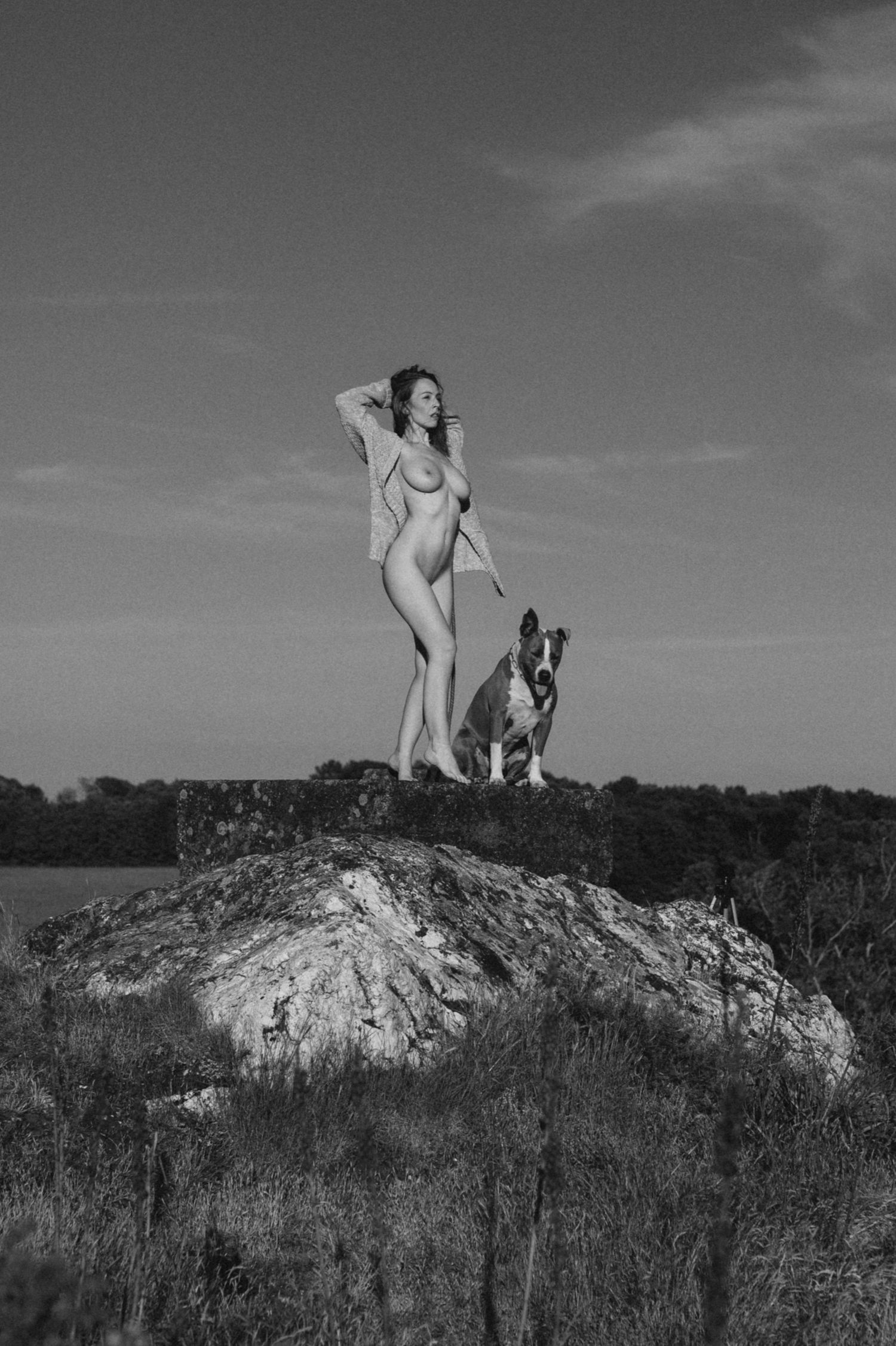 Black and white photo of a woman standing on a rock with a dog on a leash against a dramatic sky.