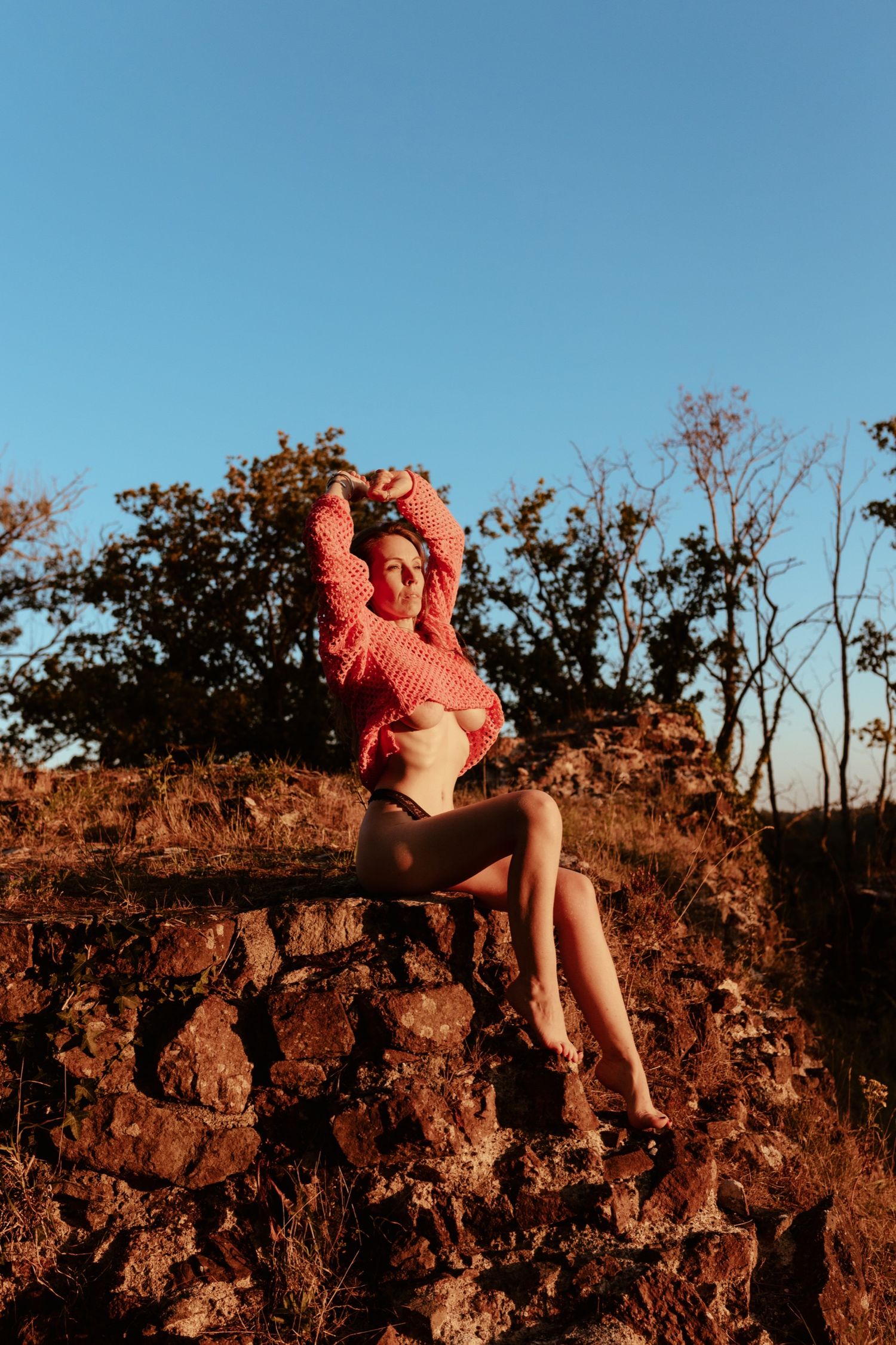 Woman in red crop top sits on stone wall at golden hour with trees and blue sky backdrop.