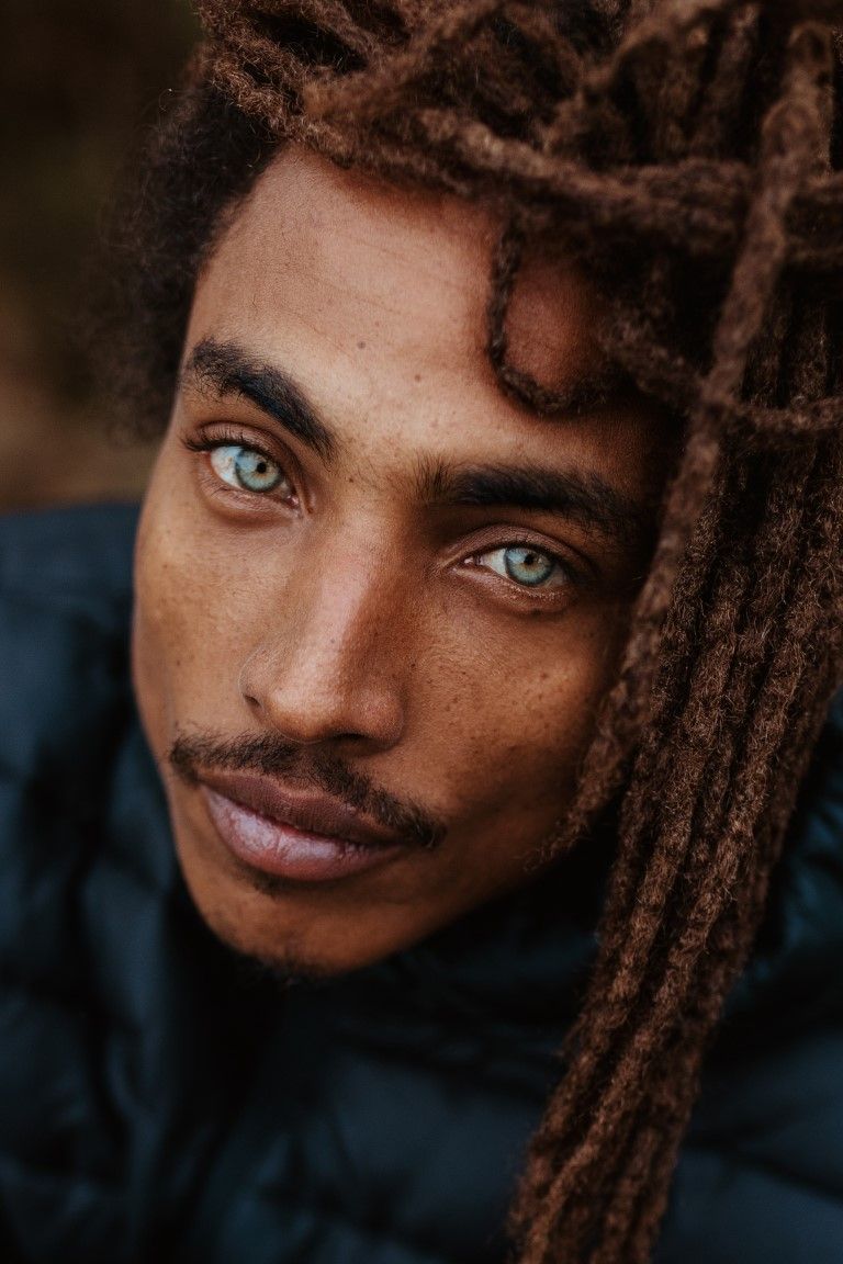Portrait en gros plan d'un homme aux longues dreadlocks brunes et aux yeux bleu-vert saisissants, vêtu d'une veste sombre.