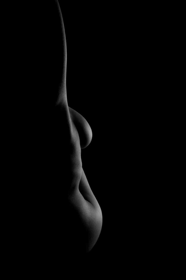 Black and white fine art nude silhouette photograph highlighting feminine body curves against dark background.