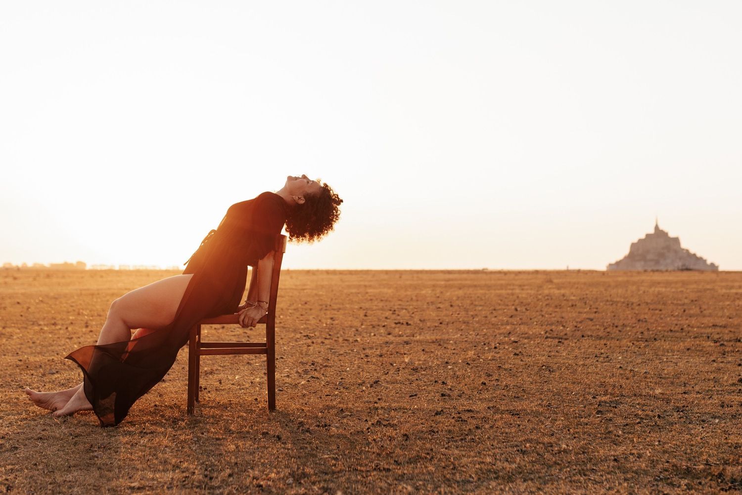 Woman in black dress leaning back on a wooden chair in a vast desert plain with Mont Saint-Michel in the background.