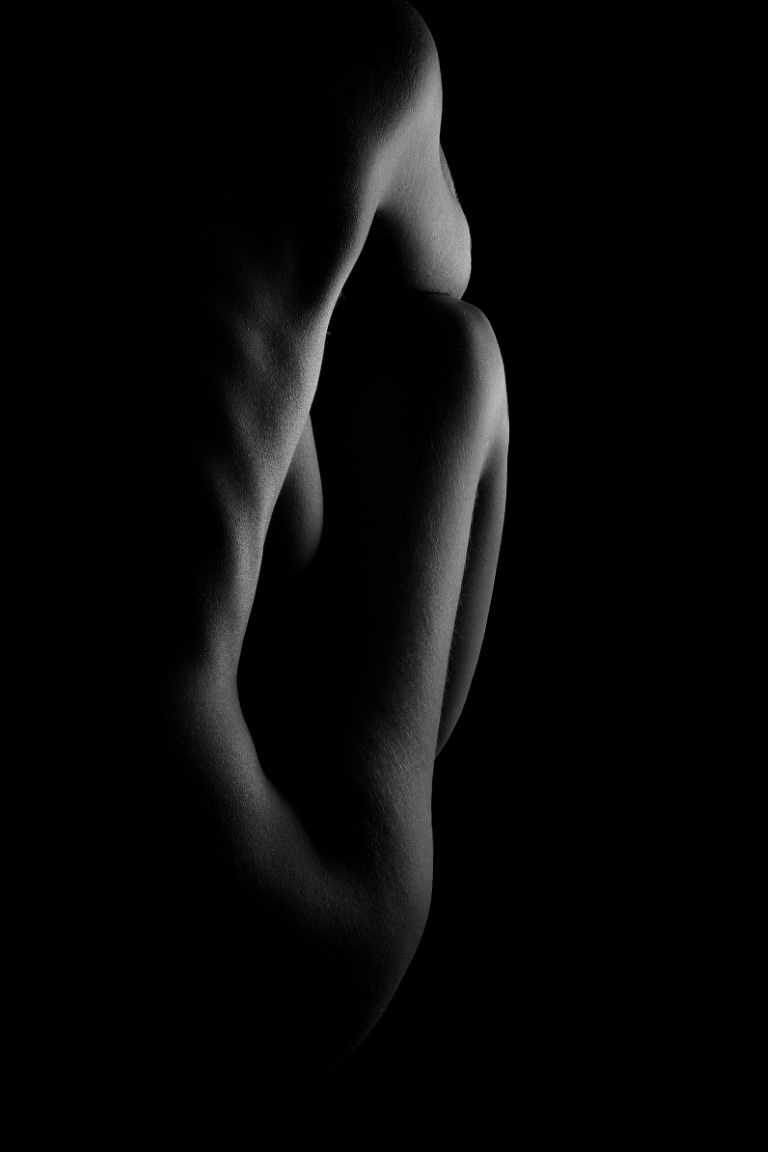 Black and white fine art nude photograph showcasing dramatic side profile with chiaroscuro lighting against dark background.